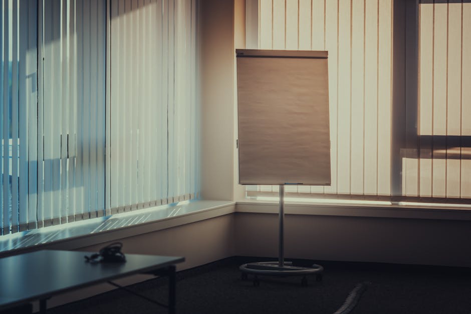 A quiet, empty meeting room featuring vertical blinds, flipchart stand, and soft lighting, perfect for business presentations.